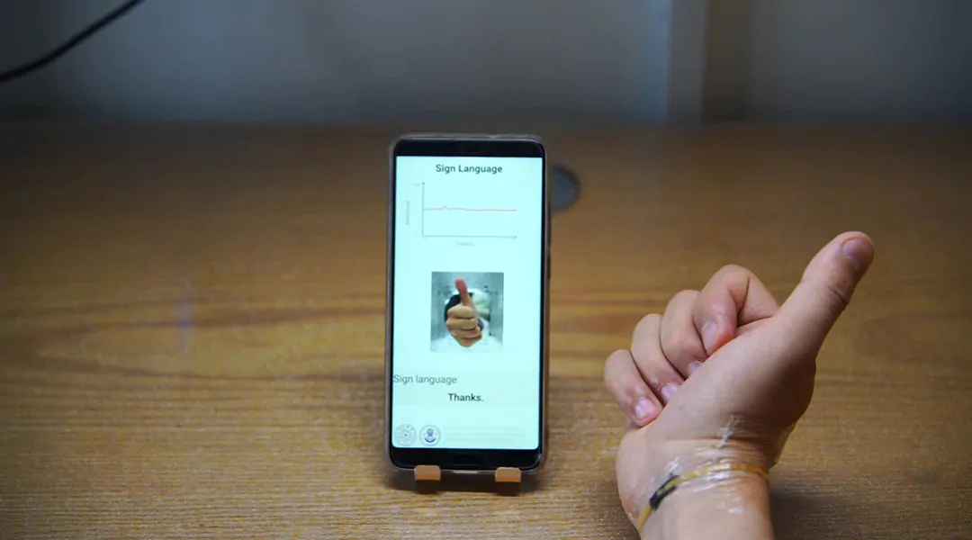 Hand on table using sign language while using a skin like sensor, a phone displays words.
