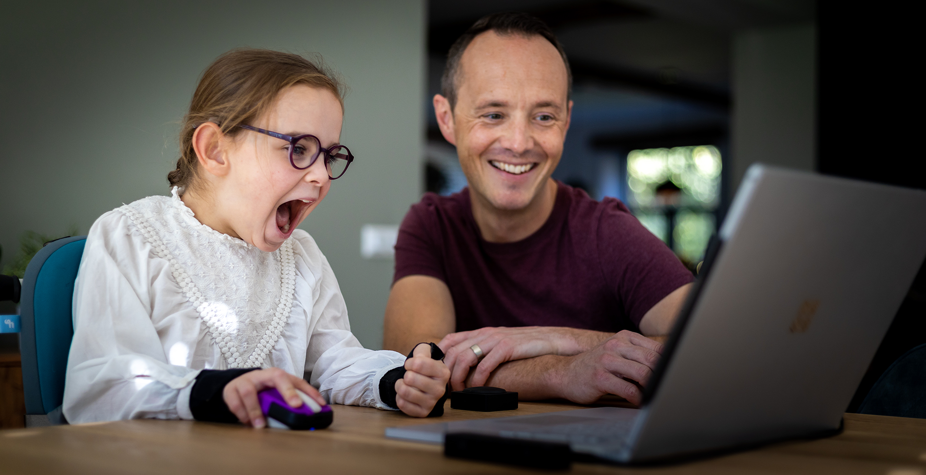 Little girl with CP using an adaptive mouse happily. Her father smiles next to her.